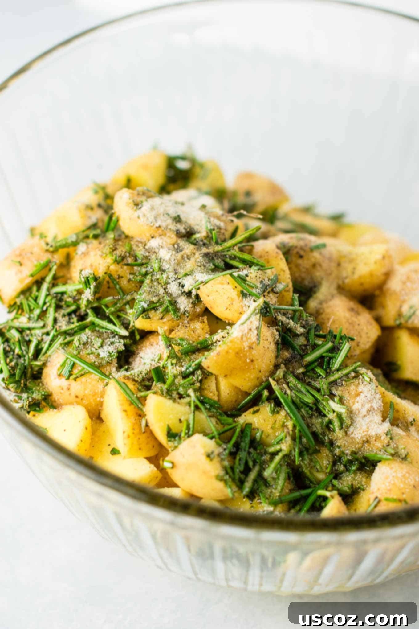 A large mixing bowl filled with raw, quartered baby potatoes, seasoned with fresh chopped rosemary, salt, pepper, garlic powder, and onion powder, ready for olive oil.