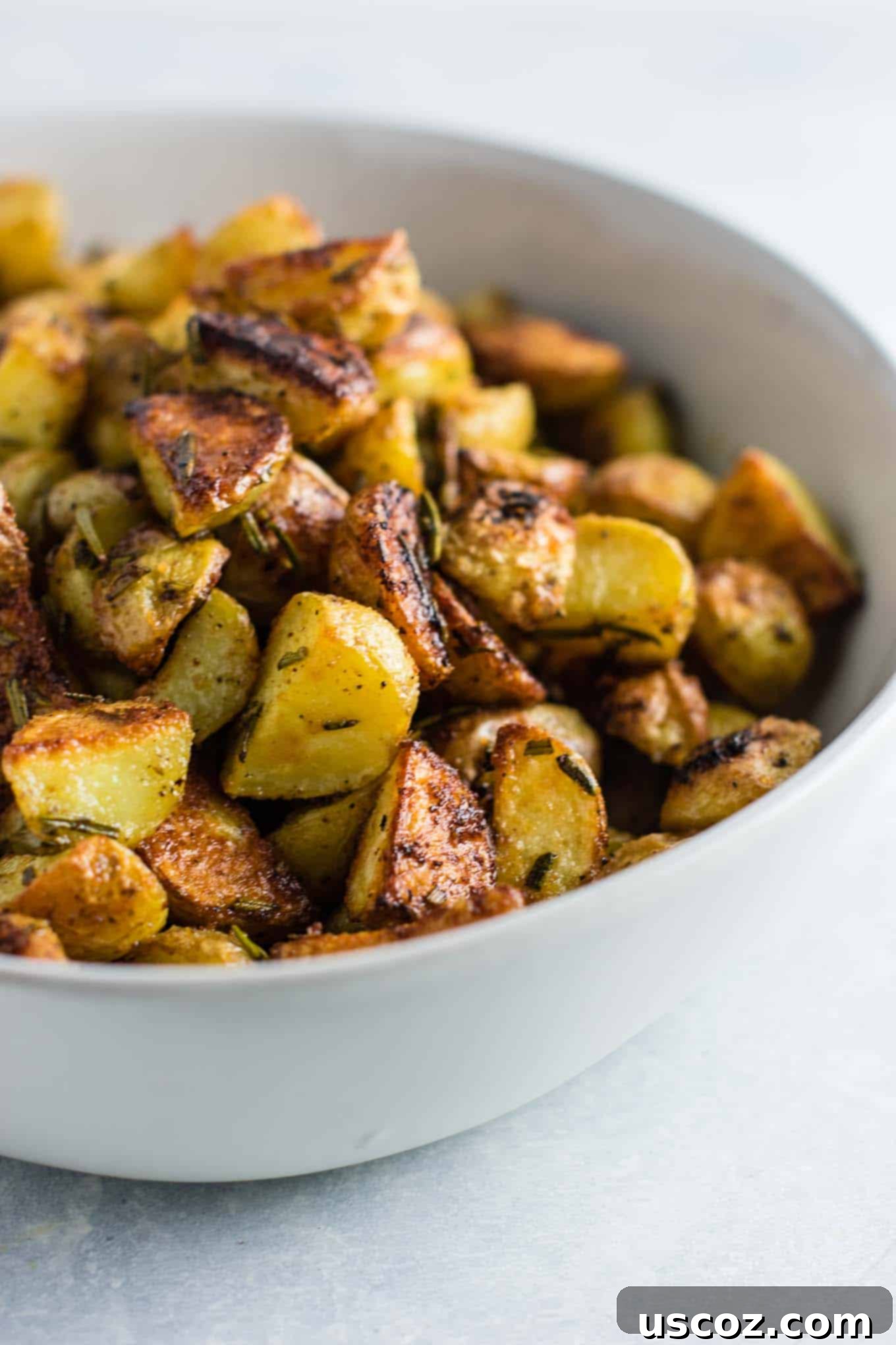 A small white bowl filled with a generous serving of crispy rosemary roasted potatoes, with a few fresh rosemary sprigs for garnish, ready to be enjoyed.