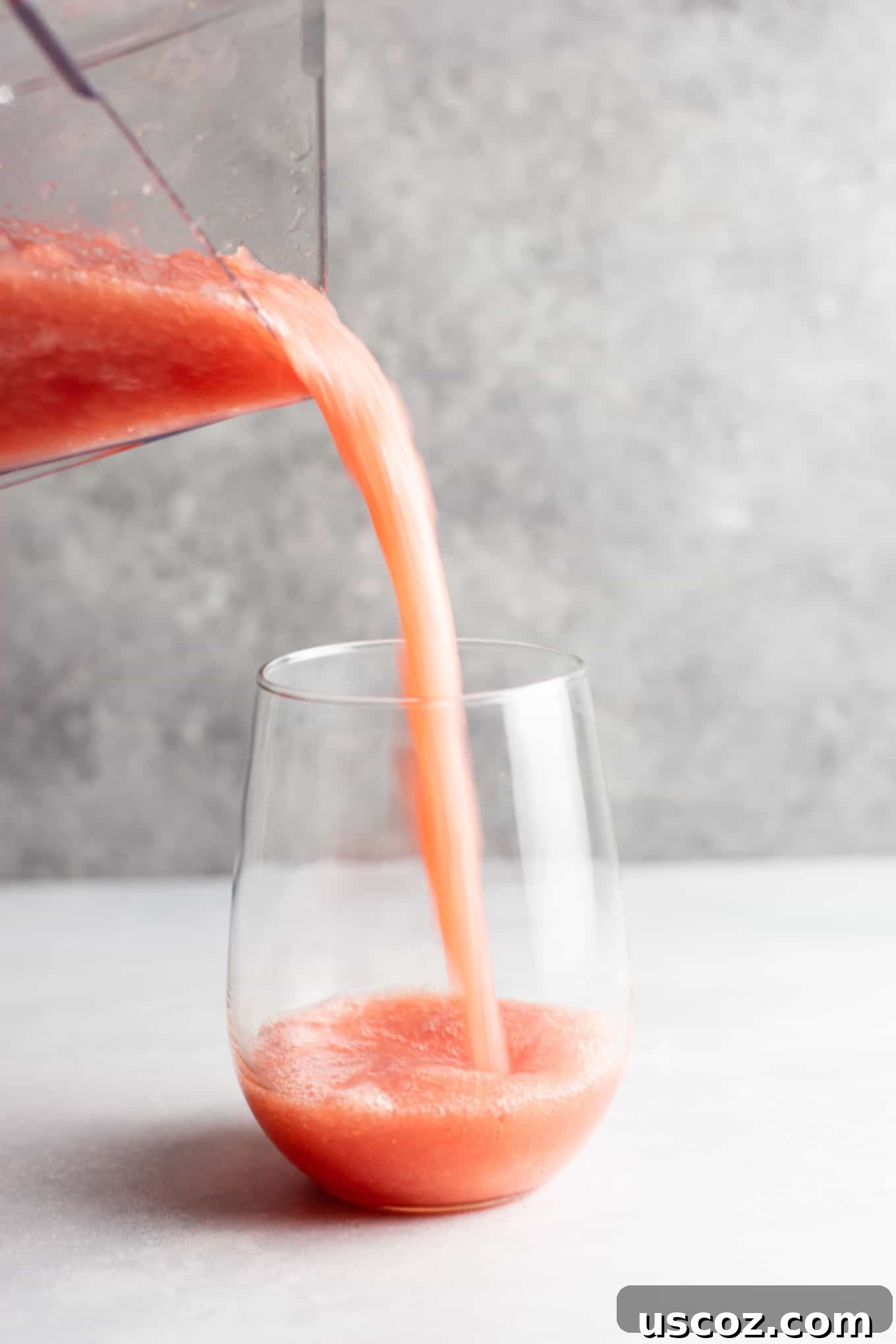 A vibrant pink watermelon white wine slushie being poured into a tall glass, showcasing its smooth, icy texture, with fresh watermelon chunks in the background.