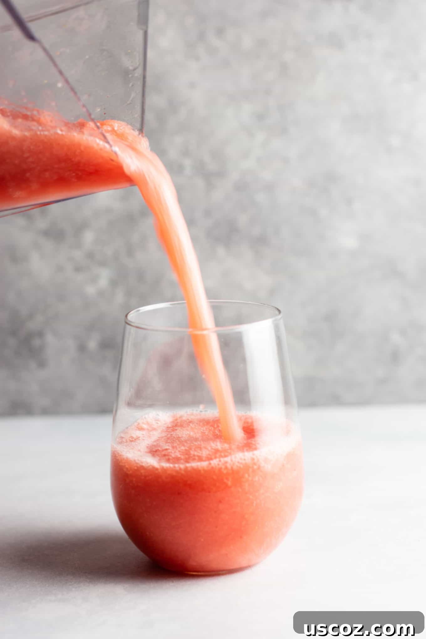 A hand holding a glass of watermelon white wine slushie, with a blurred outdoor summer background, emphasizing refreshment.