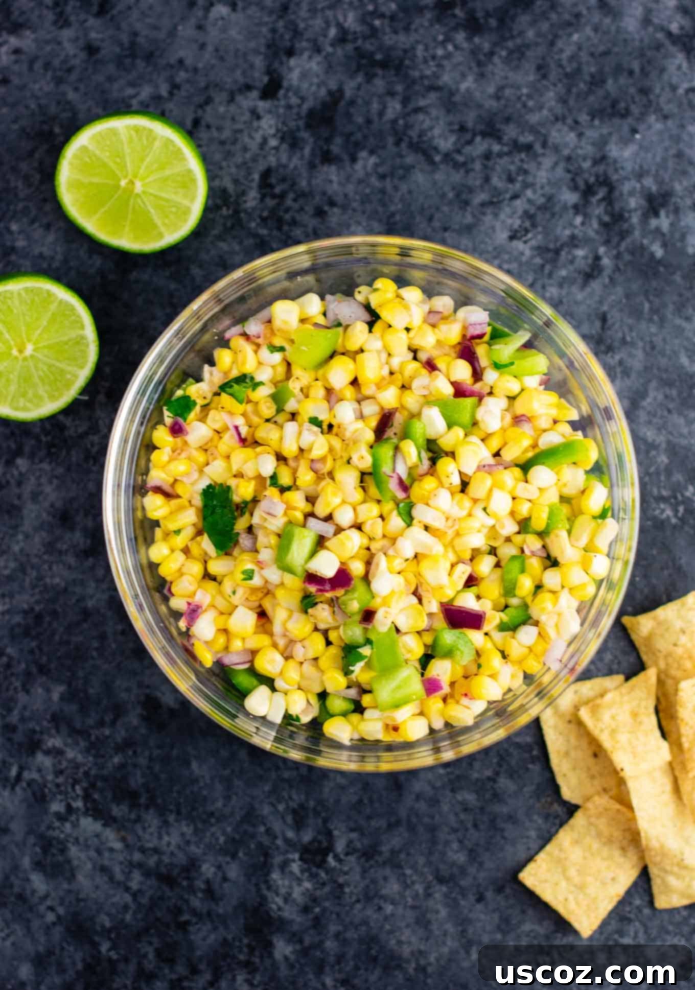 A large bowl of homemade fresh corn salsa with yellow and white corn kernels, diced red onion, and green bell pepper. Tortilla chips are arranged around the bowl, inviting a delicious dip.