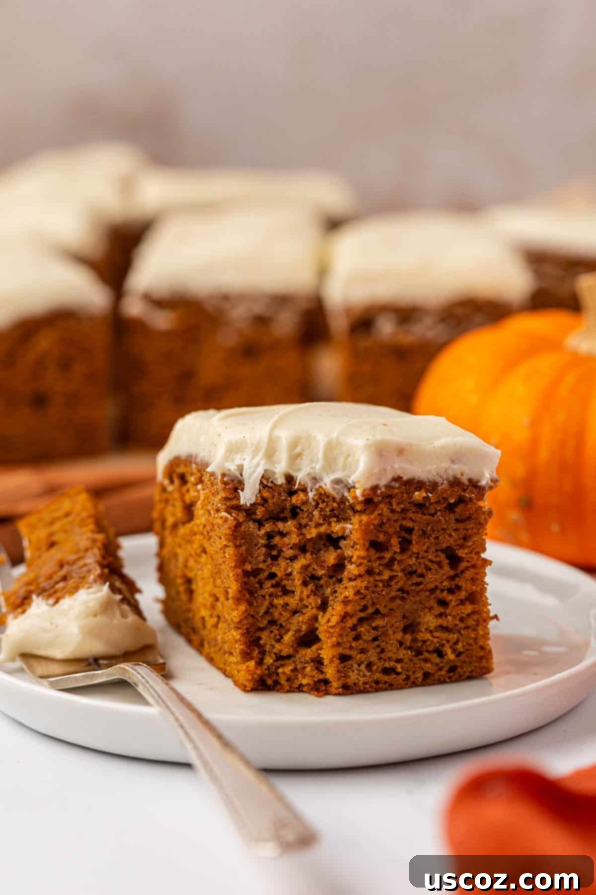 A slice of pumpkin cake with cream cheese frosting, garnished with a tiny pumpkin decoration, on a plate next to a fall-themed napkin